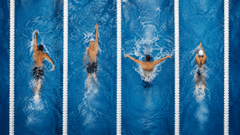 Cuatro nadadores ejecutando los 4 estilos de natación en calles paralelas de pileta competiva